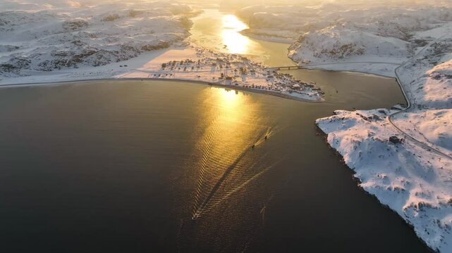 Aerial view of the Teriberka river and a snow-covered village reflecting the vibrant golden light of a winter sunset, Russia