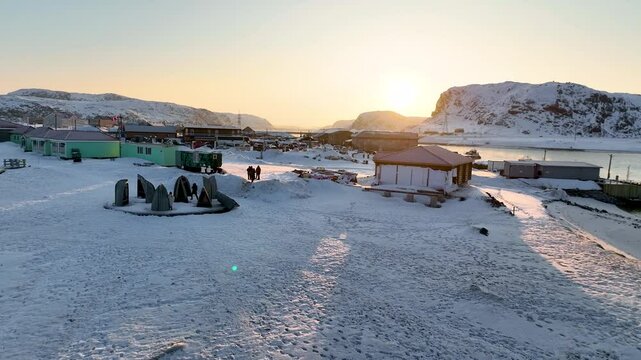 Teriberka, Russia - December 12, 2024: Aerial view of the snow-covered Teriberka village with traditional buildings, art installations, and mountains under a low winter sun, Russia