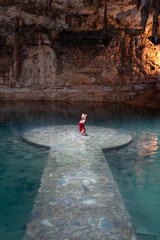 Woman standing in turquoise waters of underground cenote, raising her arms, enjoying the mystical atmosphere