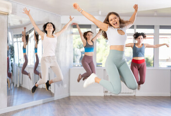 Fototapeta premium Cheerful sporty young girl captured in air while jumping during lively dance fitness routine with group of women in studio