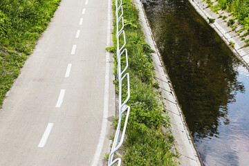 Empty bicycle road along canal. Modern cycling path with white markings. Clean pavement and safety railing. Green vegetation on both sides. Calm water reflecting trees and sky.