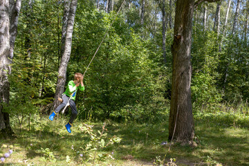 Child joyfully swings from rope tied to tree in lush forest. Outdoor recreation © Алла Чеснокова