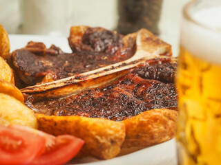 Plate with cooked high quality T bone steak, wedges and tomato in focus. Big glass of fresh lager beer out of focus in foreground. Tasty dinner with premium meat and delicious drink