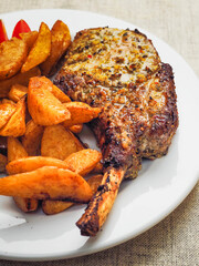 A plate of food with a premium pork chop in the bone, wedges potatoes, and tomatoes. The plate is set on a table with a fork and a knife. Fine tasty dinner with high quality meat.