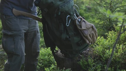 A man places his backpack on a stump in the forest and begins unpacking essential survival items for wilderness camping. Bushcraft and outdoor preparation.