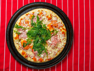 A pizza with ham and spinach on a black tray. The pizza is sitting on a red and white stripe table cloth. Fine Italian cuisine with fresh ingredients ready to be cooked. Fast food meal.