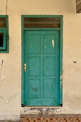 Old vintage turquoise wooden door with worn paint on a weathered beige wall, rass handle n glass transom above. Urban detail with rustic and aged character residential door, detail shot, peeling paint