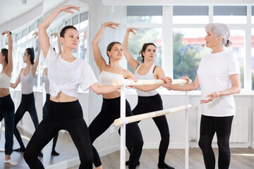 Fototapeta premium Concentrated woman practicing demi plie at barre during group ballet class for beginners under guidance of experienced elderly female instructor..