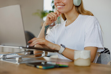 A young woman wearing headphones is diligently working on her laptop in a comfy and cozy home...