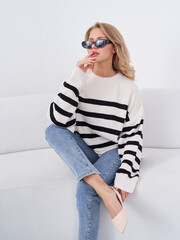 Stylish person with sunglasses sits on a white sofa wearing a black-and-white striped sweater and...