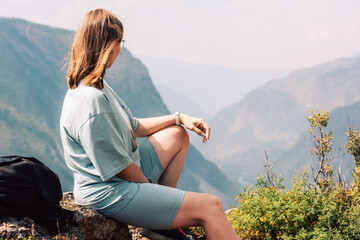Young woman in sportswear sits on a rock ledge, peacefully observing scenic mountain landscape during a summer hiking adventure.