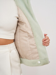 Woman wearing white top and pants holds open mint green jacket to reveal its beige lining. Hand...