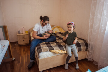 Father lovingly holds a baby on a bed while a young child enjoys a snack nearby. The cozy room features warm tones and a comfortable atmosphere, highlighting family togetherness