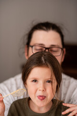 Young girl playfully sticks out her tongue while holding a lollipop. An adult with glasses is in the background, creating a joyful and relaxed atmosphere, emphasizing family fun and bonding