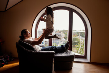 Father joyfully lifts his daughter in a warm, sunlit room with a large arched window, creating a moment of connection and happiness in a cozy home environment
