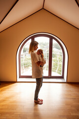 Young mother cradling her newborn baby in a sunlit room, standing beside a large arched window, creating a serene and intimate atmosphere filled with love and warmth