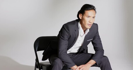Asian male model entering frame, sitting on chair in studio on cue adjusting blazer for pose - Powered by Adobe