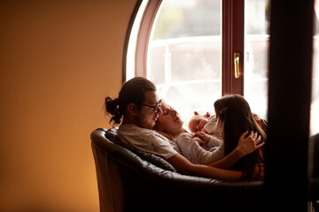 Parents and children are cuddling on a comfortable armchair near a window, enjoying a quiet moment of togetherness and creating a warm family atmosphere