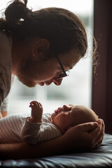 Father gently cradles his newborn baby, sharing an intimate moment by a window. The soft light highlights their connection, emphasizing the tenderness and love between them
