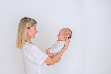 Happy mother holding her newborn baby in her arms, smiling and looking at him, standing in front of a white wall, wearing casual clothes, enjoying motherhood