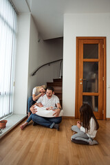 Parents sitting on the floor of a cozy living room, enjoying quality time while their newborn sleeps peacefully in a baby nest, and their older daughter plays nearby