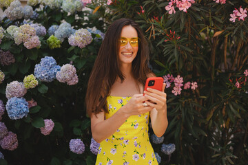 Attractive brunette long hair woman wear yellow dress and sunglasses hold mobile phone, look happy and smiling. Amazing enjoy woman walk near blossom flowers.