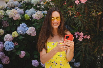 Attractive brunette long hair woman wear yellow dress and sunglasses hold mobile phone, look happy and smiling. Amazing enjoy woman walk near blossom flowers.