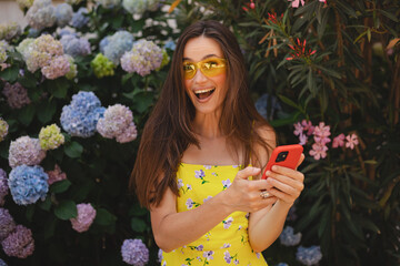 Attractive brunette long hair woman wear yellow dress and sunglasses hold mobile phone, look happy and smiling. Amazing enjoy woman walk near blossom flowers.