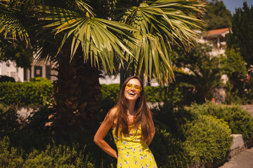 Brunette long hair tourist woman wear yellow dress and sunglasses standing near palm tree, enjoy of trip vacation. Happy girl on vacation smiling, walking. Hot summer day mood.