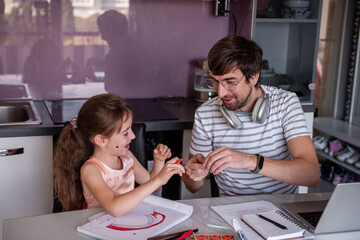 Father working from home while caring for his little daughter, guiding her as she draws with crayons at the kitchen table. Enjoying quality time together, they share laughter and creativity