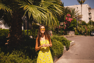Brunette long hair tourist woman wear yellow dress and sunglasses standing near palm tree, enjoy of trip vacation. Happy girl on vacation smiling, walking. Hot summer day mood.
