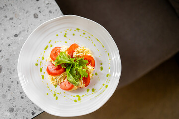 Fresh salad with sliced tomatoes, scrambled eggs, and green lettuce, garnished with green sauce, served on a white plate atop a speckled countertop. 