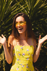 Excited young woman in yellow floral dress and yellow sunglasses smiling outdoors on a sunny day, standing in front of palm leaves with joyful expression. Here am i!