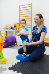 Physiotherapist assists a young boy patient wearing in special shoes in maintaining balance during a physical rehabilitation activity with fitness block
