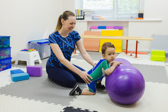 Child physical therapy session with specialist in rehab center. Little boy patient with muscle weakness doing treatment exercises standing in knees near fitness ball