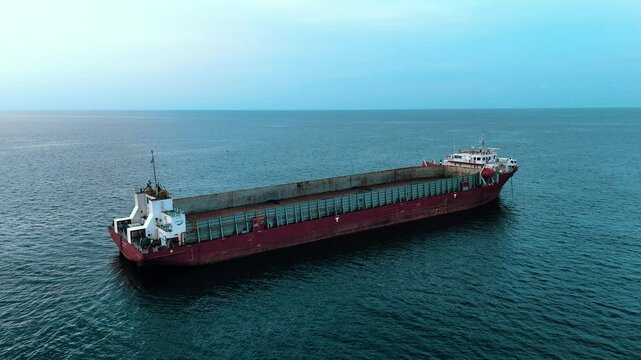 A large empty red barge or cargo ship sits alone on a calm open sea under a pale blue sky