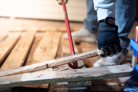 A man is forcefully prying apart a wooden pallet with a crowbar. The scene captures the intensity of manual labor, recycling, and dismantling in a raw, realistic context.