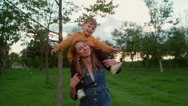 Smiling little boy rides on his mothers shoulders in a green park at sunset, enjoying playful bonding time outdoors. Young mom and her son on her shoulders looking at camera and laughing 