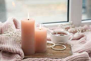 Burning candles, flowers and tray on knitted plaid near window