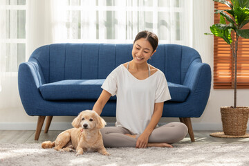 Asian smiling woman relaxing with her dog indoors, sitting on cozy carpet in front of blue sofa,...