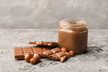 Jar of sweet chocolate spread and hazelnuts on grey background