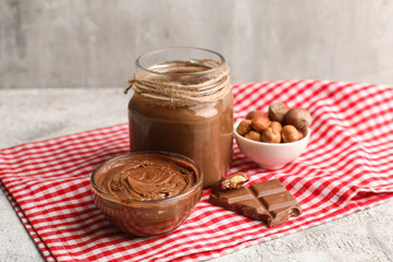 Jar and glass bowl of sweet chocolate spread with hazelnuts on grey background