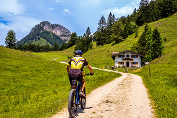 Fototapeta premium Mountain biker rides along a scenic dirt path near a rustic house in lush green countryside during daylight