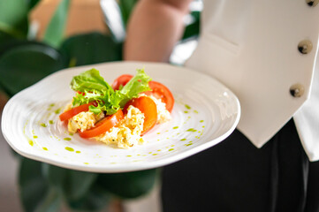 A close-up of a fresh salad with scrambled eggs, sliced tomatoes, and green lettuce on a white plate, garnished with green sauce. The dish is held by a person in a white shirt