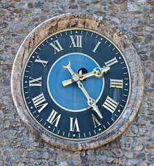 A large stone-framed clock with golden Roman numerals and ornate hands. Set in a rough stone wall, its dark blue face contrasts with the shimmering metal and aged, weathered texture.