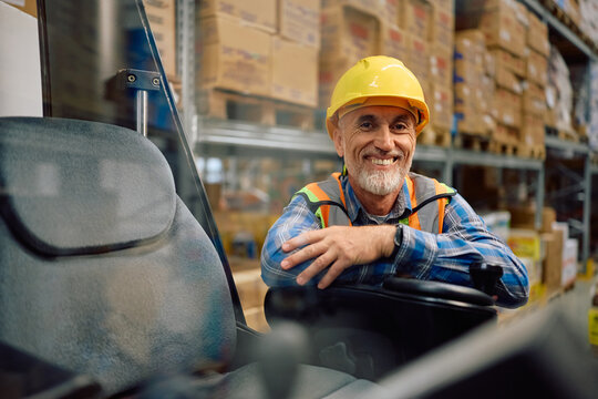 Happy senior worker operating forklift in warehouse and looking at camera.