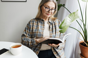 Smart senior caucasian lady in glasses reading classic or scientific book while having cup of tea in morning, spending her leisure time sitting at home at table next to window and big houseplan