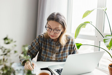 Retired people and remote education. Upper angle indoor view of intelligent senior caucasian woman in glasses writing brief summary in copybook while study online at home using laptop