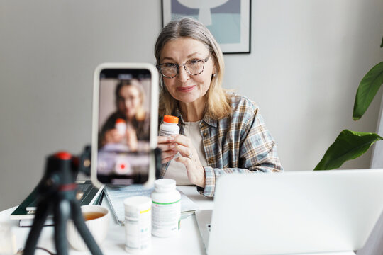 Indoor image with selective focus on aged woman nutritionist holding bottle of food supplement and recording video for social media, sharing her knowledge and giving advice on how to stay healthy