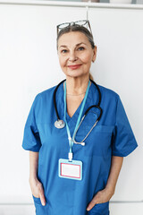 Vertical indoor portrait of senior female doctor in blue uniform with stethoscope and name tag, standing with hands in pocket and smiling at camera after successful work day at hospital